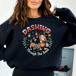 May include: Black sweatshirt with a festive graphic. The design features a cartoon dog wearing a Santa hat and antlers, decorated with Christmas lights, and the text "Dashing Through The Snow" in a circular design.
