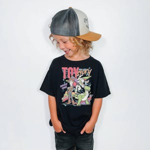 May include: A black t-shirt with a colourful graphic of "Toy Story" characters and the text "Toy Story" and "Since 1995". The child is also wearing a grey and tan trucker hat.