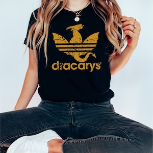 Pu&ograve; includere: Maglietta nera con una grafica di drago dorata usurata e il testo "dracarys" in oro.