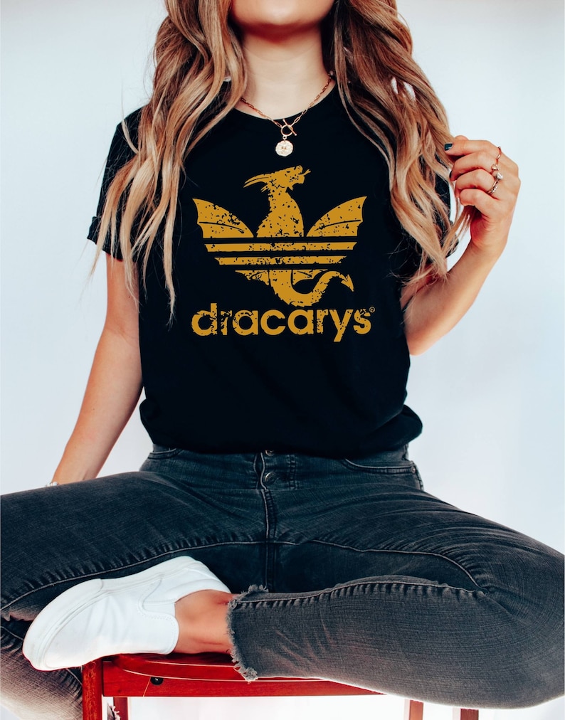 Pu&ograve; includere: Maglietta nera con una grafica di drago dorata usurata e il testo "dracarys" in oro.