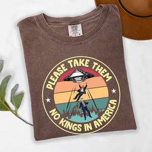 Op de afbeelding: Bruin Comfort Colors T-shirt met een cirkelvormige afbeelding met een retro zonsondergang, een UFO en twee figuren die worden ontvoerd. De tekst luidt "PLEASE TAKE THEM" en "NO KINGS IN AMERICA".
