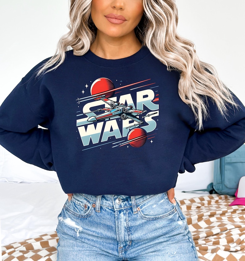 Puede incluir: Una sudadera azul marino con un gr&aacute;fico de una nave espacial volando por el espacio con el texto "Star Wars" en letras blancas y rojas.