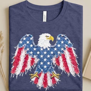 May include: A navy blue t-shirt featuring a central graphic of a bald eagle with a distressed, frayed-edge appearance. The eagle's body is patterned with white stars on a blue background, while its wings and tail display red and white stripes.
