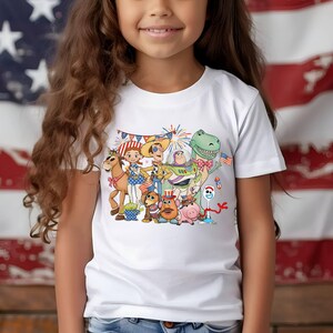 Puede incluir: Una camiseta blanca de manga corta con un estampado gr&aacute;fico colorido de varios personajes animados, incluyendo un vaquero, un guardi&aacute;n espacial, un dinosaurio y un cerdo. Los personajes est&aacute;n decorados con detalles patri&oacute;ticos en rojo, blanco y azul.