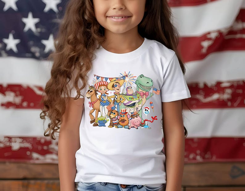 Puede incluir: Una camiseta blanca de manga corta con un estampado gr&aacute;fico colorido de varios personajes animados, incluyendo un vaquero, un guardi&aacute;n espacial, un dinosaurio y un cerdo. Los personajes est&aacute;n decorados con detalles patri&oacute;ticos en rojo, blanco y azul.