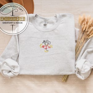 May include: A gray sweatshirt with a simple embroidered design of Mickey Mouse in yellow, red, and black. The sweatshirt has a crew neck and long sleeves.