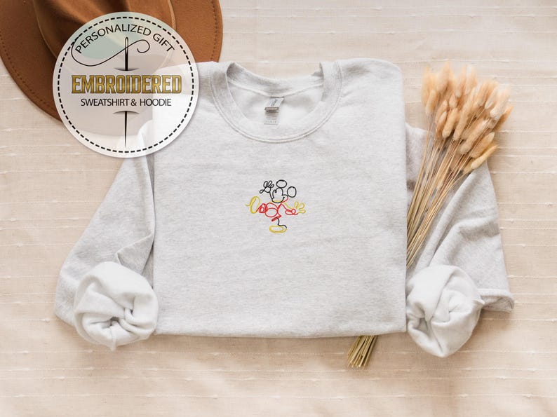 May include: A gray sweatshirt with a simple embroidered design of Mickey Mouse in yellow, red, and black. The sweatshirt has a crew neck and long sleeves.