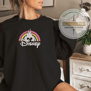 May include: Black sweatshirt with a rainbow and a white castle graphic with the word "Disney" underneath. The sweatshirt is being worn by a person.