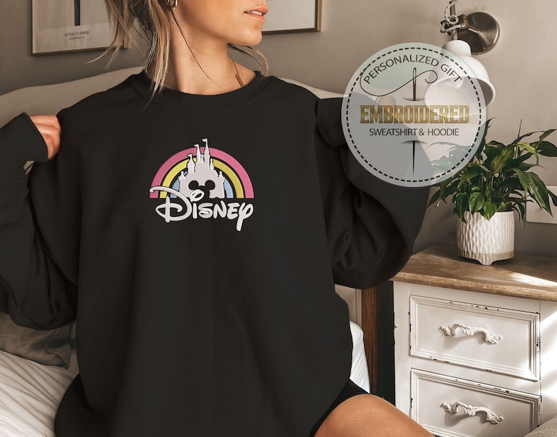 May include: Black sweatshirt with a rainbow and a white castle graphic with the word "Disney" underneath. The sweatshirt is being worn by a person.