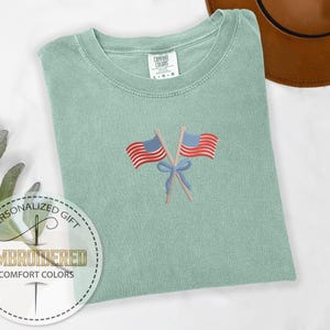 May include: A light green Comfort Colors t-shirt with an embroidered design of two American flags crossed with a blue bow. The shirt has a crew neck and short sleeves. The shirt is a personalized gift.