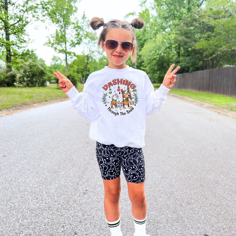 May include: A young person wearing a white sweatshirt with the text "Dashing Through The Snow" and a graphic of reindeer and holiday decorations. They are also wearing black biker shorts with a smiley face pattern.