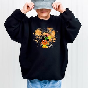 May include: Black sweatshirt featuring a colorful cartoon character design. The design includes a character with orange, yellow, and green accents, set against a gold splatter background. The sweatshirt is paired with light blue jeans and a gray baseball cap.