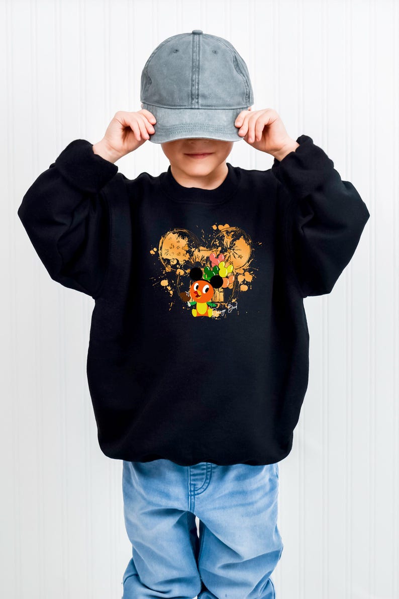 May include: Black sweatshirt featuring a colorful cartoon character design. The design includes a character with orange, yellow, and green accents, set against a gold splatter background. The sweatshirt is paired with light blue jeans and a gray baseball cap.