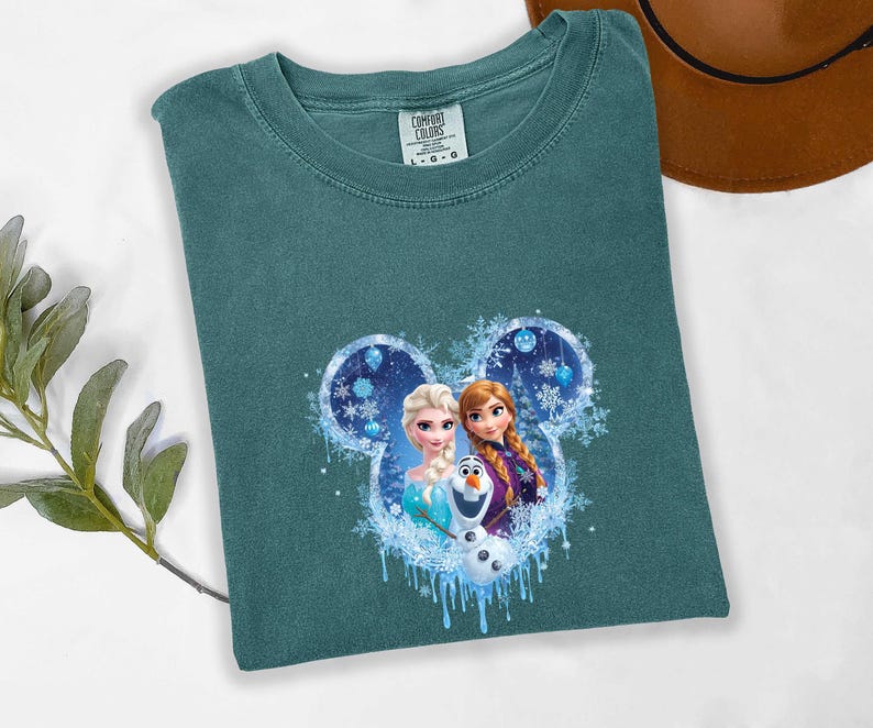 May include: Teal Comfort Colors t-shirt featuring a Disney Frozen graphic. The graphic includes Elsa, Anna, and Olaf in a Mickey Mouse-shaped design with snowflakes and blue accents. The shirt is laid flat.
