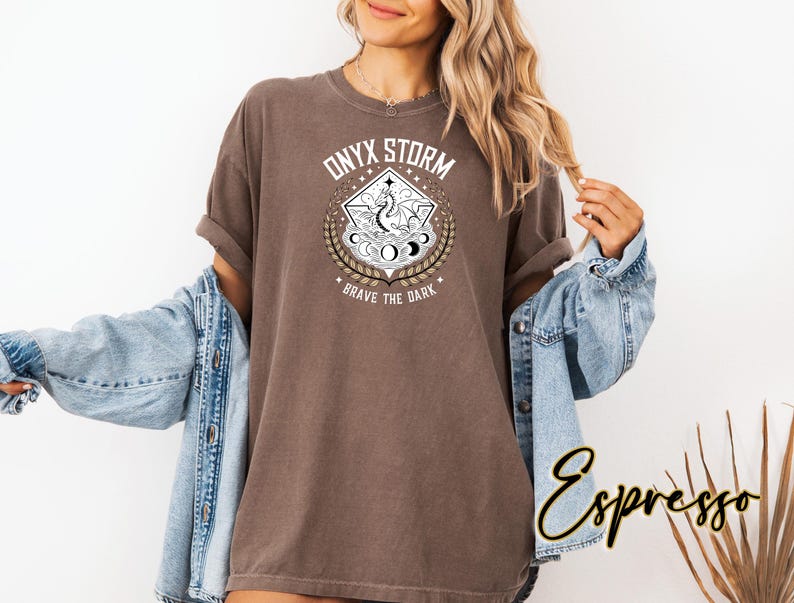 May include: A brown t-shirt with a white graphic design featuring a dragon, moons, and stars. The graphic is surrounded by a diamond shape with the text "ONYX STORM" and "BRAVE THE DARK".