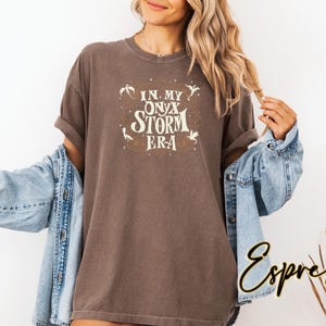 May include: A brown t-shirt with the text "In My Onyx Storm Era" in a vintage style font. The text is surrounded by small, stylized dragon illustrations.