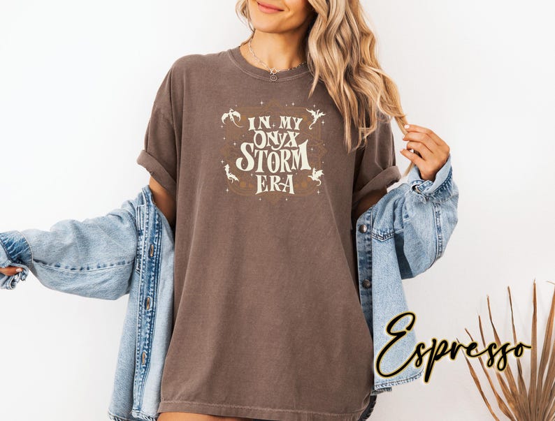 May include: A brown t-shirt with the text "In My Onyx Storm Era" in a vintage style font. The text is surrounded by small, stylized dragon illustrations.