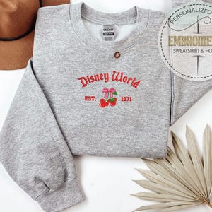 May include: A heather gray sweatshirt with the embroidered text "Disney World" in red script, along with a bow and cherries graphic. The year "1971" is below the graphic. A brown hat and a dried palm leaf are in the background.