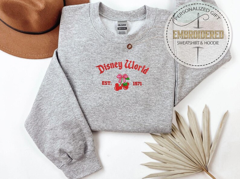 May include: A heather gray sweatshirt with the embroidered text "Disney World" in red script, along with a bow and cherries graphic. The year "1971" is below the graphic. A brown hat and a dried palm leaf are in the background.