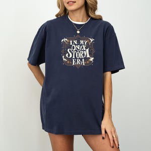 May include: A navy blue t-shirt with a graphic print of a stylized design that says "In My Onyx Storm Era" with dragons and stars.