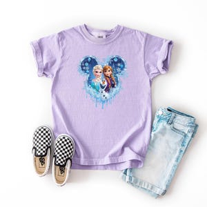 May include: A lavender t-shirt featuring a Disney-themed graphic of Elsa and Anna from Frozen, set within a Mickey Mouse-shaped design. The image includes light blue denim shorts and black and white checkered slip-on shoes.