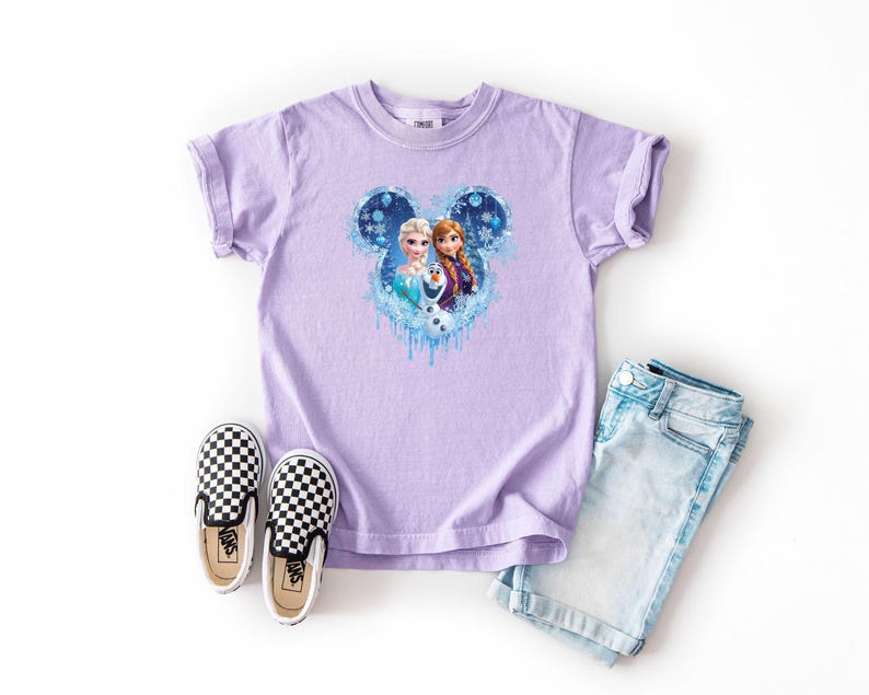 May include: A lavender t-shirt featuring a Disney-themed graphic of Elsa and Anna from Frozen, set within a Mickey Mouse-shaped design. The image includes light blue denim shorts and black and white checkered slip-on shoes.