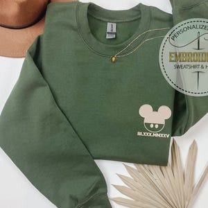 May include: An olive green sweatshirt with a round neckline, featuring a light beige cartoon mouse design and Roman numerals. A gold necklace with a pendant is draped on the sweatshirt. The image also includes a brown hat and a decorative tag.