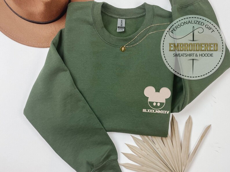 May include: An olive green sweatshirt with a round neckline, featuring a light beige cartoon mouse design and Roman numerals. A gold necklace with a pendant is draped on the sweatshirt. The image also includes a brown hat and a decorative tag.