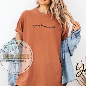 May include: A rust-colored t-shirt with the embroidered text "in my Disney mom era". The shirt has rolled-up sleeves and a relaxed fit. A circular logo with the words "Personalized Gift" and "Embroidered Comfort Colors" is visible.