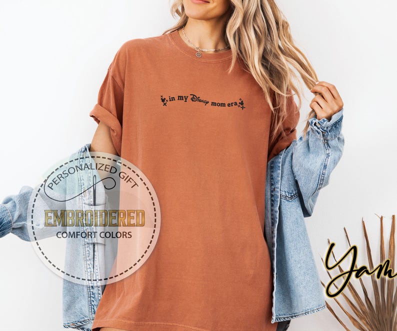 May include: A rust-colored t-shirt with the embroidered text "in my Disney mom era". The shirt has rolled-up sleeves and a relaxed fit. A circular logo with the words "Personalized Gift" and "Embroidered Comfort Colors" is visible.