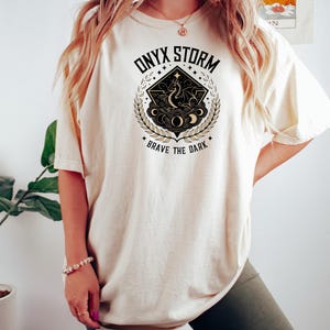 May include: A cream colored t-shirt with a graphic design of a dragon in a circle with the text "ONYX STORM" and "BRAVE THE DARK" in black and gold lettering.