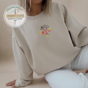 May include: A beige sweatshirt with a small embroidered image of Mickey Mouse in black, red, and yellow thread. The sweatshirt has a crew neck and long sleeves.