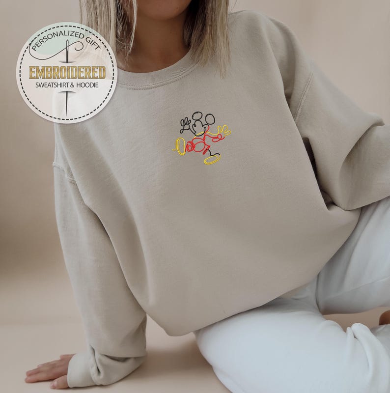 May include: A beige sweatshirt with a small embroidered image of Mickey Mouse in black, red, and yellow thread. The sweatshirt has a crew neck and long sleeves.