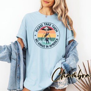 Op de afbeelding: Lichtblauw T-shirt met een cirkelvormige afbeelding. De afbeelding toont een UFO, twee figuren die worden ontvoerd en de tekst "PLEASE TAKE THEM NO KINGS IN AMERICA". Het ontwerp bevat een achtergrond die op een zonsondergang lijkt.