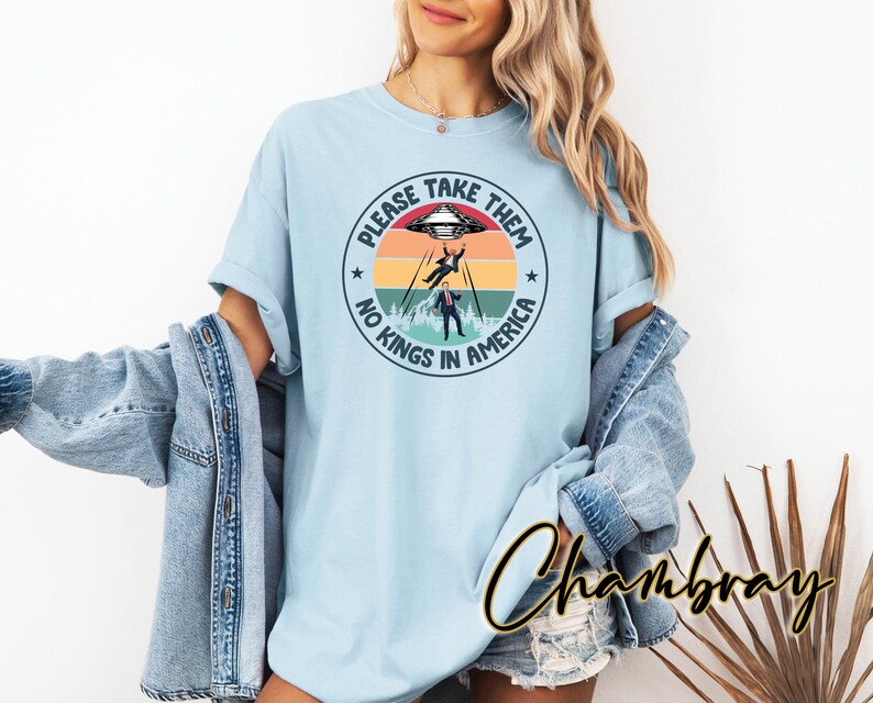Op de afbeelding: Lichtblauw T-shirt met een cirkelvormige afbeelding. De afbeelding toont een UFO, twee figuren die worden ontvoerd en de tekst "PLEASE TAKE THEM NO KINGS IN AMERICA". Het ontwerp bevat een achtergrond die op een zonsondergang lijkt.