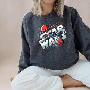 Puede incluir: Una sudadera gris oscuro con un estampado gr&aacute;fico de una nave espacial volando por el espacio con el texto "STAR WARS" en letras blancas y rojas.