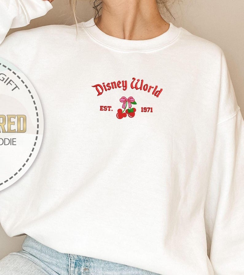 May include: White crewneck sweatshirt with the text "Disney World" in red script, along with a red bow and cherries graphic. The text "EST. 1971" is also included. The sweatshirt is a casual style.