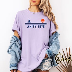 May include: A lilac-colored t-shirt with a nautical design featuring a sailboat, horizontal lines, and a setting sun. The text "AMITY 1975" is printed below the lines. The shirt is part of the "Orchid" collection.