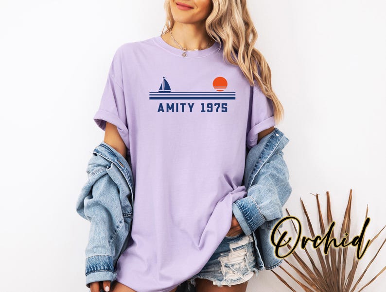 May include: A lilac-colored t-shirt with a nautical design featuring a sailboat, horizontal lines, and a setting sun. The text "AMITY 1975" is printed below the lines. The shirt is part of the "Orchid" collection.