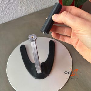 May include: A black and silver 3D printed stand for displaying small objects. The stand has a round base and a curved arm that can be adjusted to different heights. The stand is made of two pieces, a base and a removable arm.