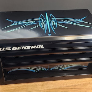 Pode incluir: Caixa de ferramentas preta U.S. GENERAL com três gavetas. A parte superior e as frentes das gavetas apresentam um design de riscas personalizado em tons de azul e verde-azulado. A caixa de ferramentas está sobre uma superfície de madeira.