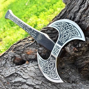 May include: A double-headed axe with a black wooden handle and silver blades. The blades are decorated with intricate Celtic knotwork designs. The axe is resting on a tree trunk.