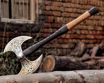 May include: A double-headed axe with a black wooden handle wrapped in brown twine. The axe heads are silver with gold Celtic knot designs. The axe is resting on a brown log.
