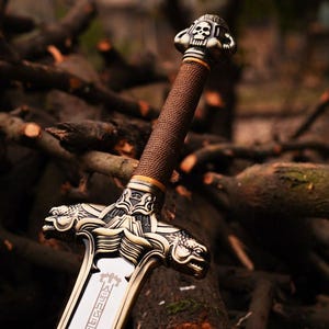 May include: A detailed close-up of a decorative sword. The hilt is wrapped in brown material, with a gold-colored metal guard featuring ornate designs and a skull at the pommel. The blade has a silver finish with etched details.