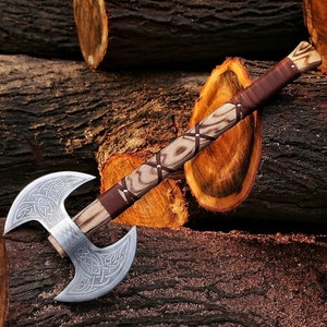 May include: A double-headed axe with a brown leather wrapped handle and a wood shaft. The axe head has intricate knotwork designs and is made of polished steel. The axe is resting on a pile of logs.