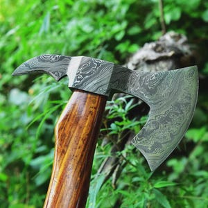 Könnte beinhalten: Eine handgefertigte Axt mit einer gemusterten Stahlklinge und einem gebogenen Haken. Der Axtkopf hat ein einzigartiges Design, und der Holzgriff hat eine satte, braune Oberfläche. Der Hintergrund ist eine verschwommene Ansicht von grünem Laub.