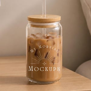May include: A clear glass tumbler with a bamboo lid and a glass straw, filled with iced coffee. The glass features a white graphic design with sunburst rays and the text "MY MORNING MOCKUPS" printed on the front surface of the container.