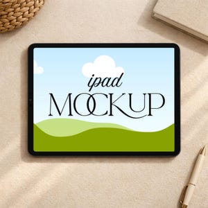 May include: A black iPad mockup displays a digital landscape with a blue sky, white clouds, and green hills. The words "ipad MOCKUP" are written in a stylish font. A pen and a book are in the background.