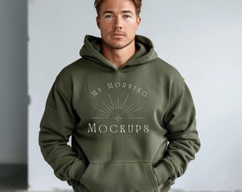 May include: An olive green hoodie with a front pocket and a hood. The hoodie features the text "MY MORNING MOCKUPS" in a white, stylized font. The model is wearing the hoodie and has their hands in the front pocket.
