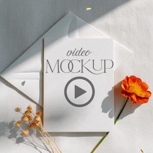 May include: A white rectangular card featuring the text video MOCKUP and a play button icon, placed atop a white envelope. A single bright orange flower is positioned to the right, and a small cluster of dried golden flowers sits to the left.